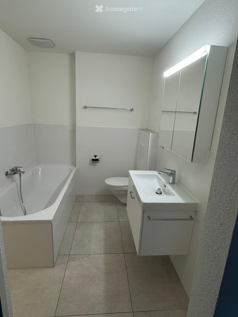 Modern bathroom with white tiled walls, a bathtub, toilet, sink, and a mirrored cabinet with built-in lighting.
