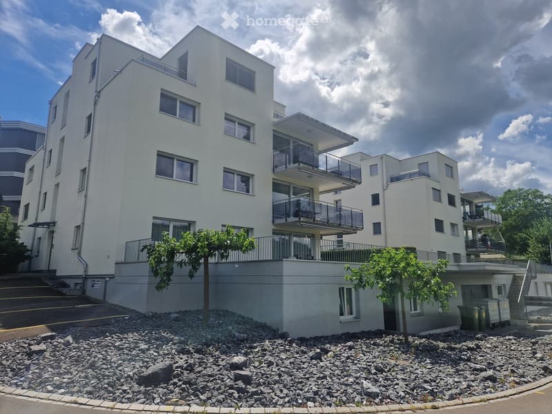 Modern multi-story residential building with white facade, large windows, and balconies. The ground floor has a raised entrance area with gravel landscaping and small trees.