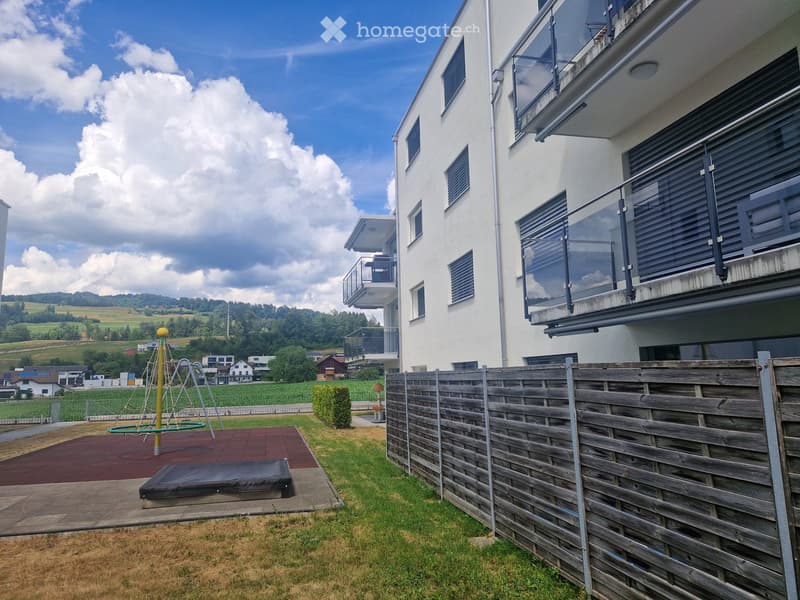 Outdoor area with a wooden fence, grassy lawn, playground equipment, and a modern building with balconies in the background.