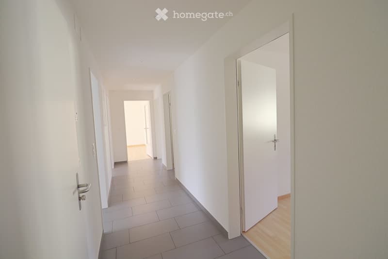 A clean, modern hallway with white walls, tiled floor, and several closed white doors. The hallway has recessed ceiling lighting and a wooden floor visible at the end of the corridor.