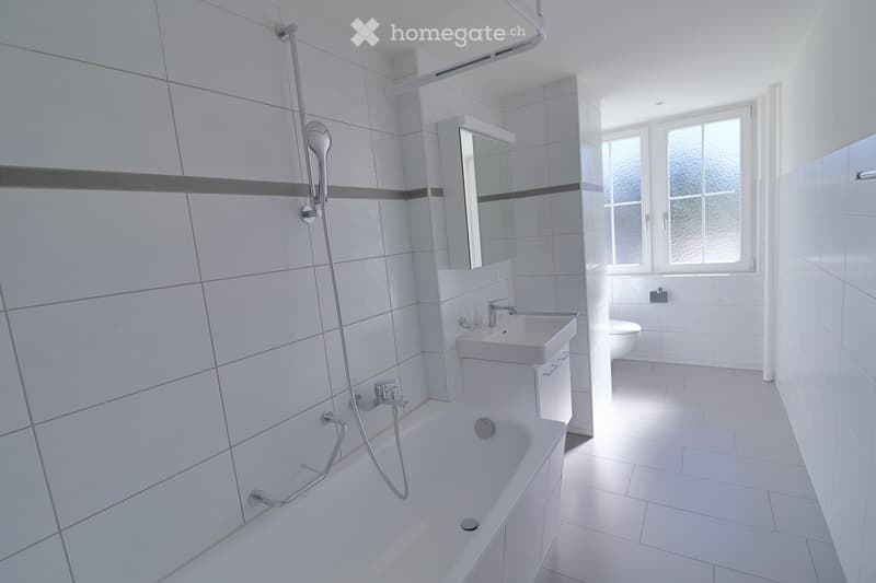 Modern bathroom with white tiles, bathtub, sink, mirror, and toilet. Features a showerhead and a window providing natural light.