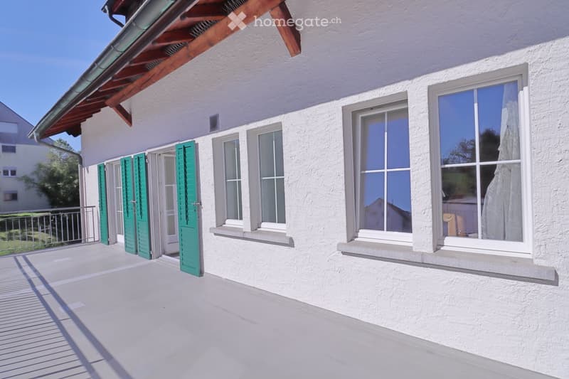 Balcony with white concrete floor, green shutters, and large windows overlooking the house exterior.