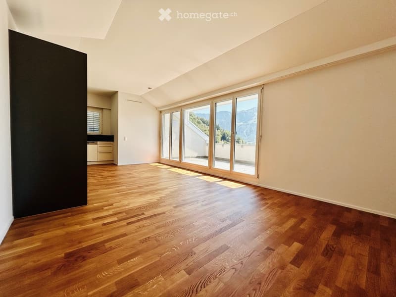 Empty room with wooden flooring, large windows offering a mountain view, and a small adjacent kitchen area.