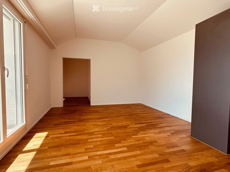 Empty room with wooden flooring, white walls, and large windows allowing natural light to enter. The room features a simple, clean design with no furniture or decorations.