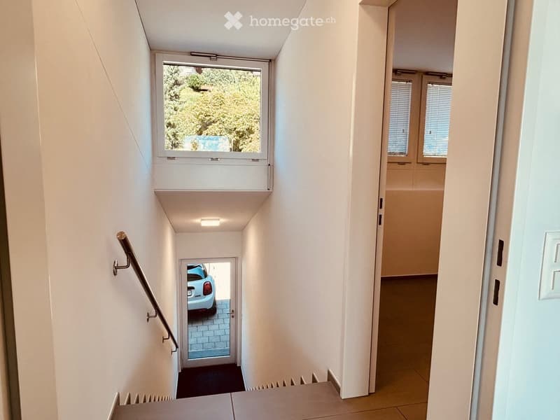 Modern hallway with white walls, a staircase leading to a parking area, and a window overlooking greenery.