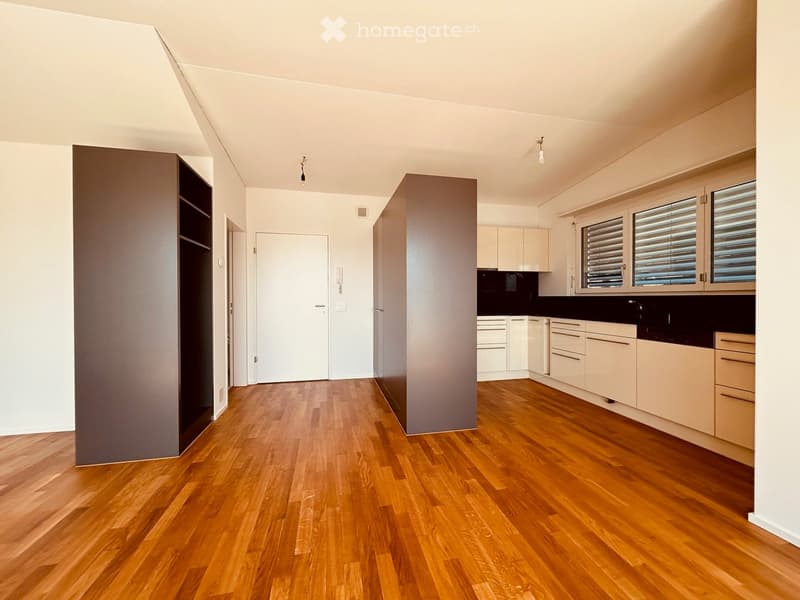 Open-plan unfurnished space with a kitchen area and living area, featuring light wooden flooring, white cabinetry, and large windows with blinds.