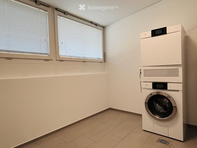 A small, clean bathroom with tiled floors, white walls, and two windows with blinds. A white washing machine and dryer are stacked against the wall.