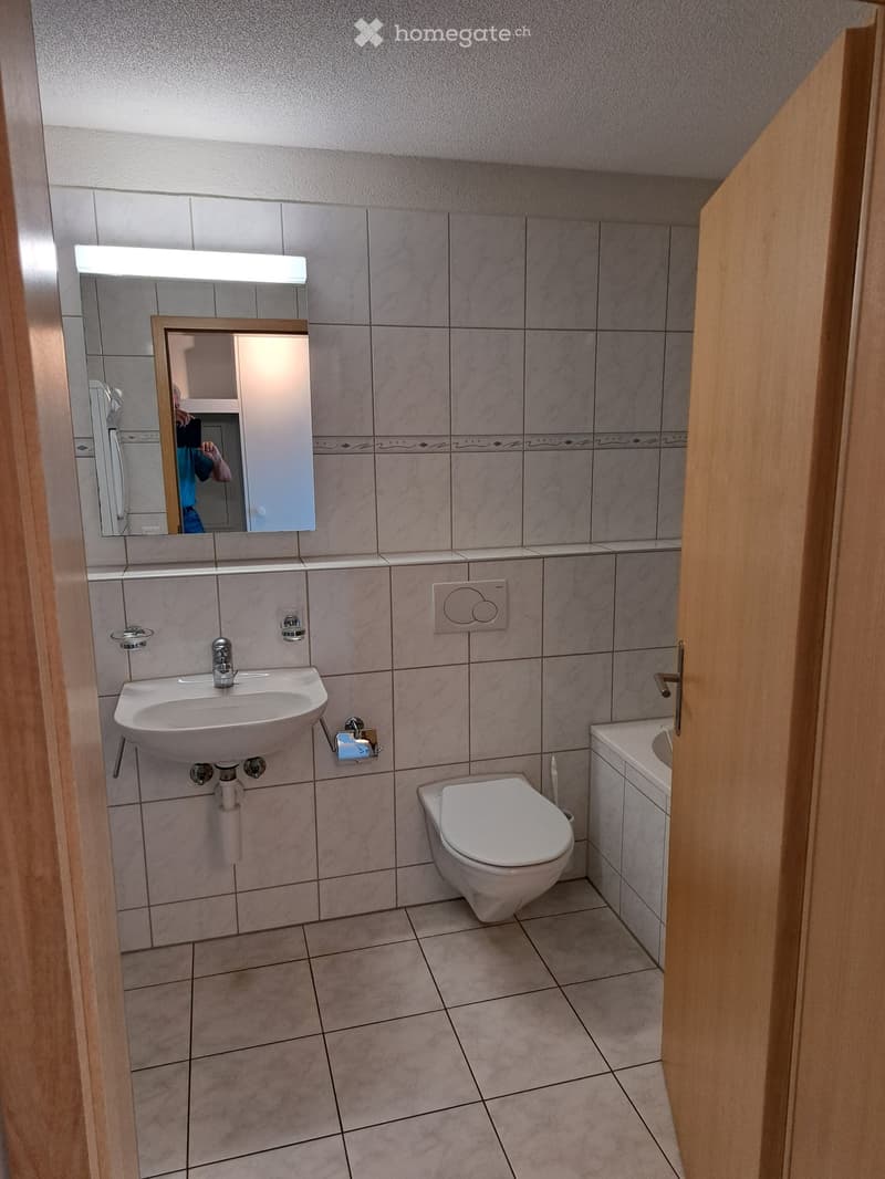 Modern bathroom with a white sink, toilet, and mirror. The walls are tiled in a light grey color with a decorative border. The floor is tiled in a light beige color.