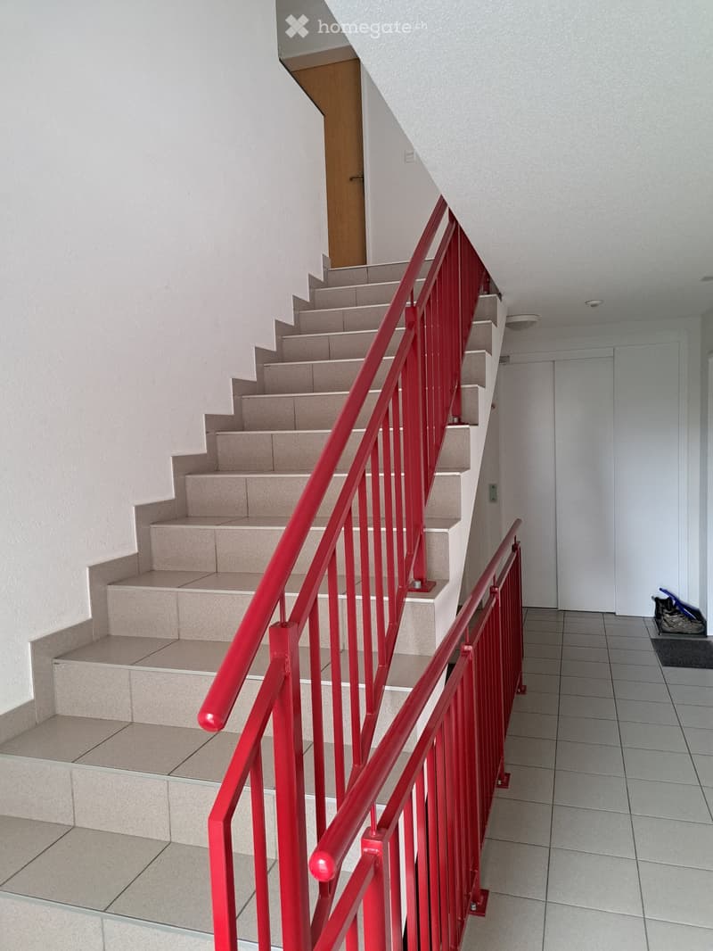 A modern staircase with red metal railings and beige tiled steps, leading upwards with white walls and a closed door at the end of the hallway.