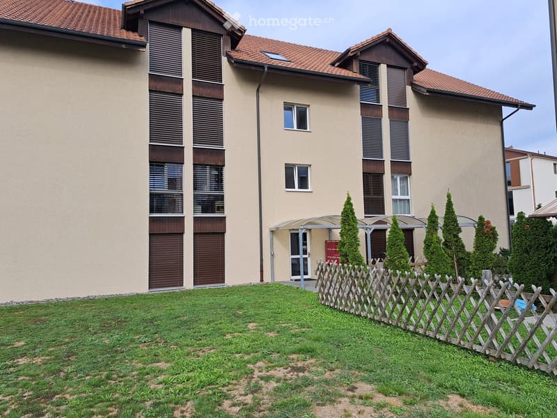 Two-story house with beige walls, brown shutters, and a red-tiled roof. The front yard features a wooden fence, neatly trimmed grass, and several tall, slender evergreen trees.