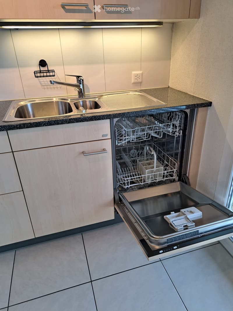 Modern kitchen with stainless steel sink, faucet, and open dishwasher showing racks and interior. Light wood cabinets and granite countertop.