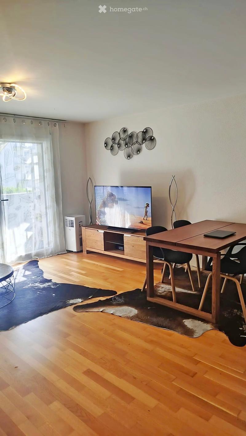A modern living room with wooden flooring, a large television, a dining table with chairs, and decorative lamps.