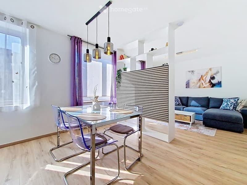Modern open-plan living and dining area with a grey sectional sofa, coffee table, glass dining table with four chairs, pendant lights, and built-in shelving unit.