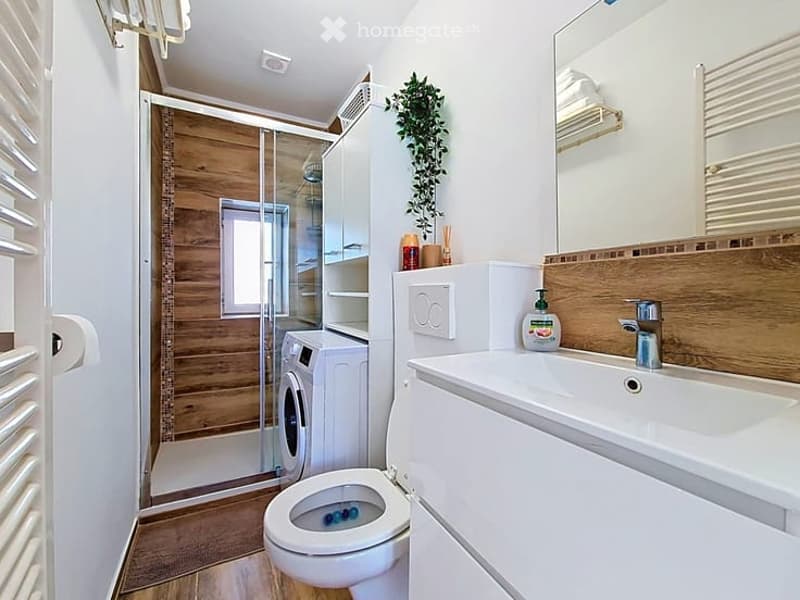 Modern bathroom with white tiles, wooden accents, a toilet, sink, mirror, and a washing machine in the corner. A small plant adds a touch of greenery.
