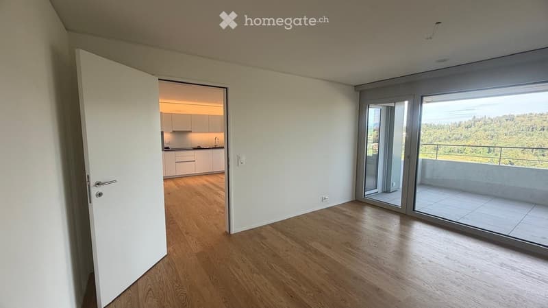 Empty room with wooden flooring, large sliding glass doors leading to a balcony with scenic view, and an open doorway to a modern kitchen.