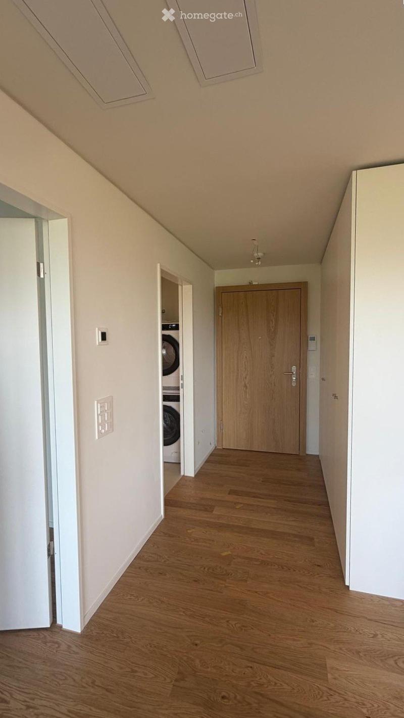 Empty hallway with wooden flooring, white walls, and a visible laundry area behind an open door.