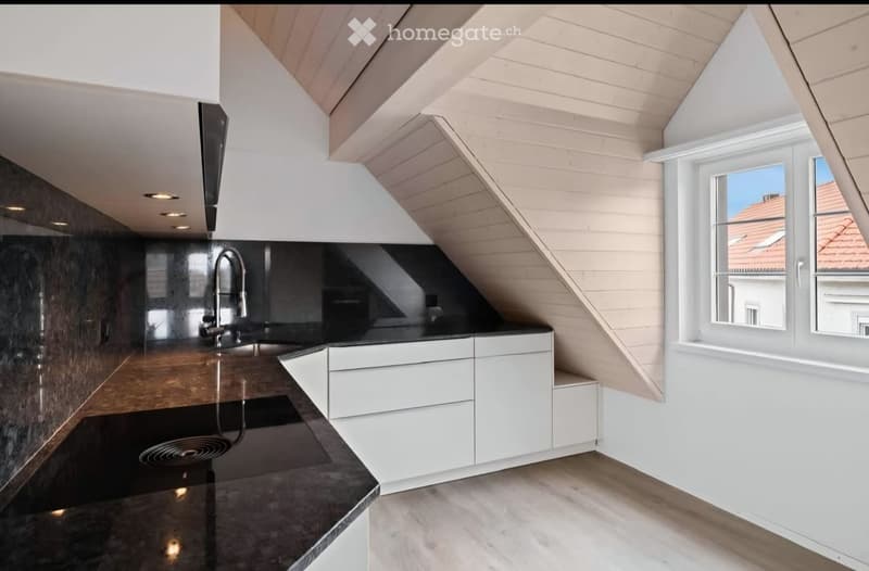 Modern kitchen with white cabinets, dark countertops, and a sleek faucet, featuring a slanted wooden ceiling and large windows.