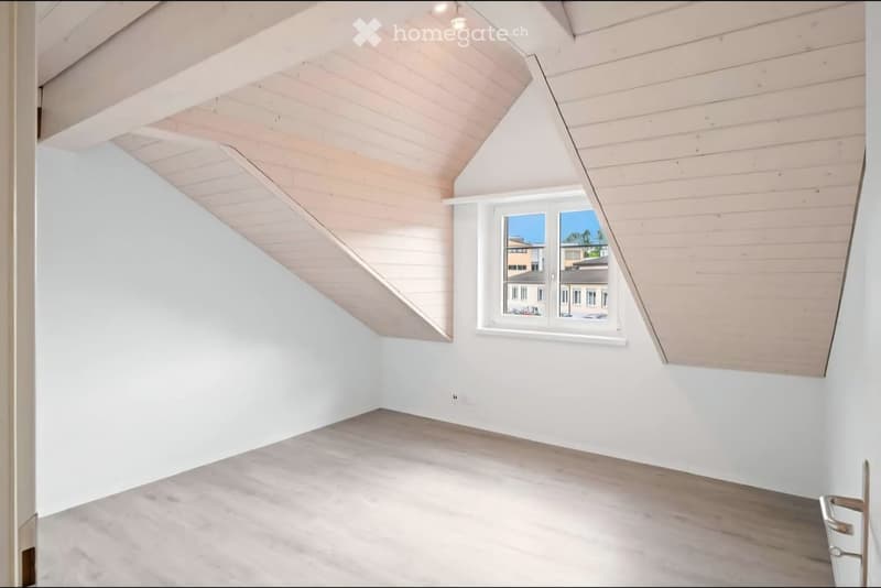Empty room with white walls, wooden floor, and a large window showing a view of neighboring buildings.