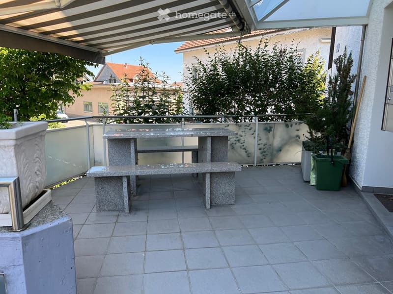 Balcony with tiled floor, stone table, glass railing, and greenery in the background.