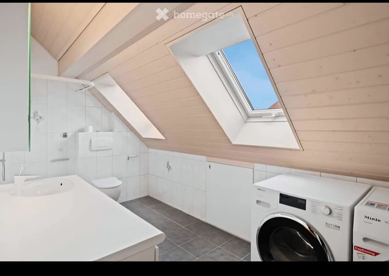 Modern bathroom with white tiles, wooden slanted ceiling, skylight window, sink, toilet, and washing machine.