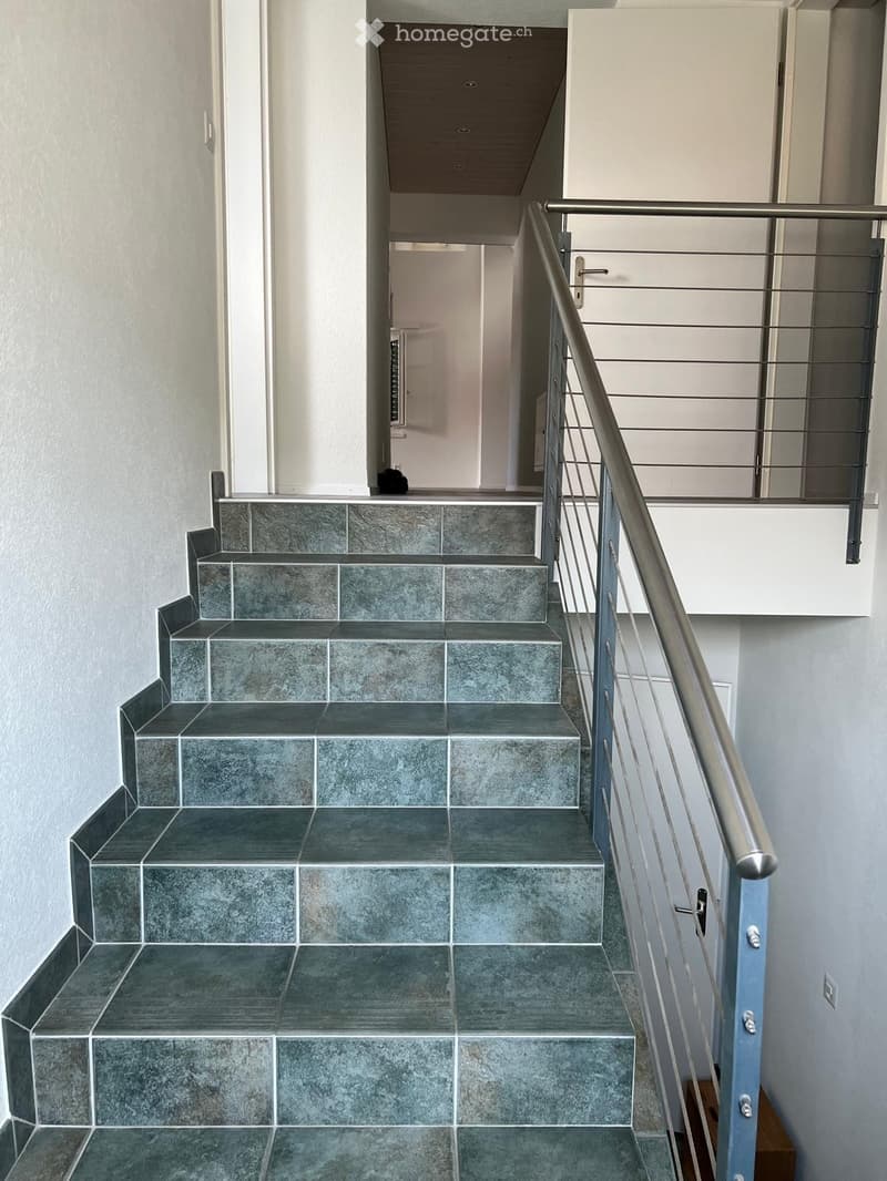 Modern staircase with grey tiled steps and a metal railing, leading to an open area with white walls and a visible door at the end.