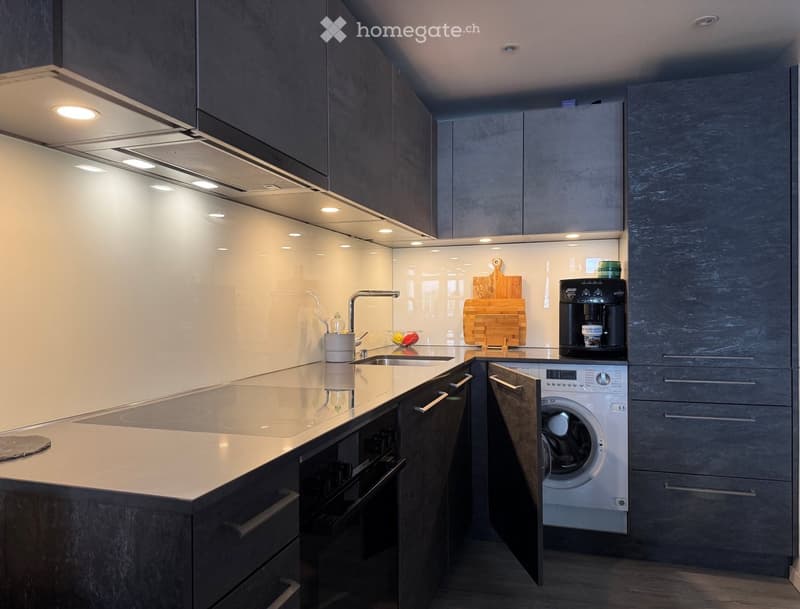 Modern kitchen with dark grey cabinets, white countertops, built-in appliances including a coffee maker, cutting board, and washing machine, and sleek lighting.