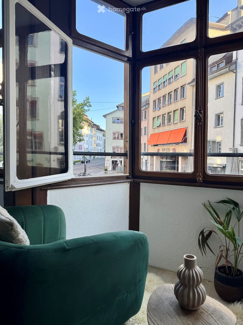 A furnished balcony with a green sofa, a small round table with a vase, and a potted plant, overlooking a city street with buildings and a clear blue sky.