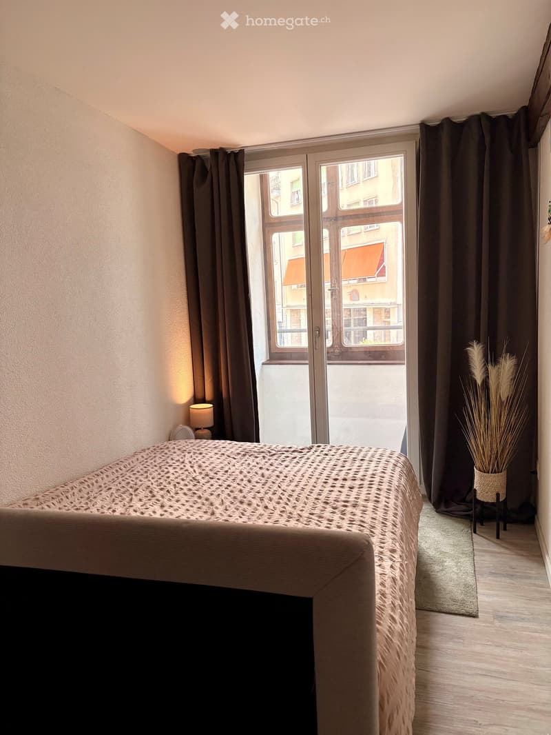 Modern bedroom with a large bed, bedside lamp, dark curtains, and a decorative vase with dried plants near the window.
