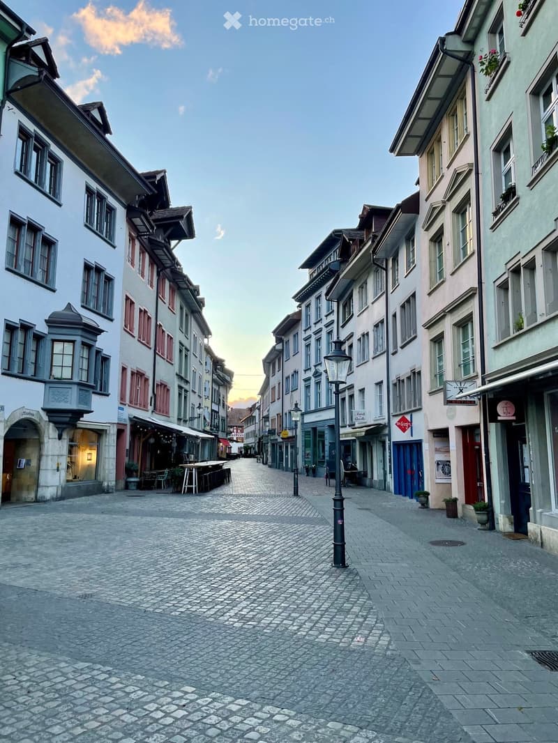 A narrow cobblestone street lined with historic buildings, featuring a mix of architectural styles, street lamps, and outdoor seating areas.