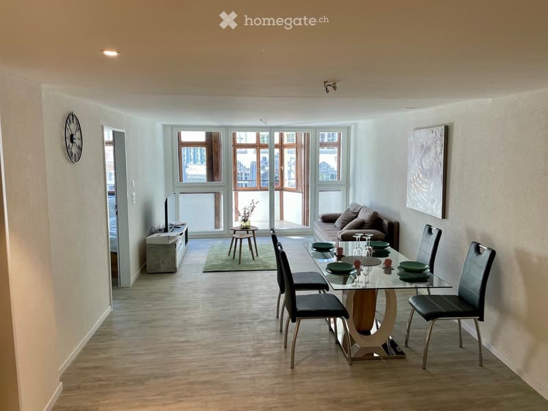 Open-plan living and dining area with a sofa, dining table, chairs, a small stool, a television, and a clock on the wall.
