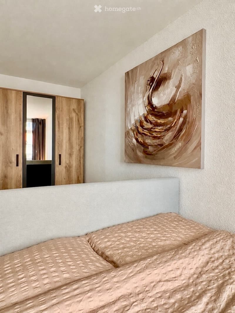 Modern bedroom with a large bed, beige bedding, a wooden wardrobe with a mirror, and an abstract painting on the wall.