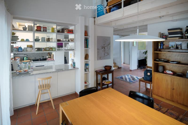 Open-plan kitchen and dining area with white cabinets, wooden table, chairs, and various kitchen items on shelves and counters.