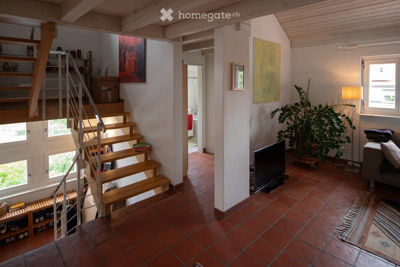 A modern, open-plan living area with a wooden staircase leading to an upper level. The space features a grey sofa, a flat-screen TV, a floor lamp, a large potted plant, and colorful artwork on the walls. The floor is tiled in terracotta, and there is a sm