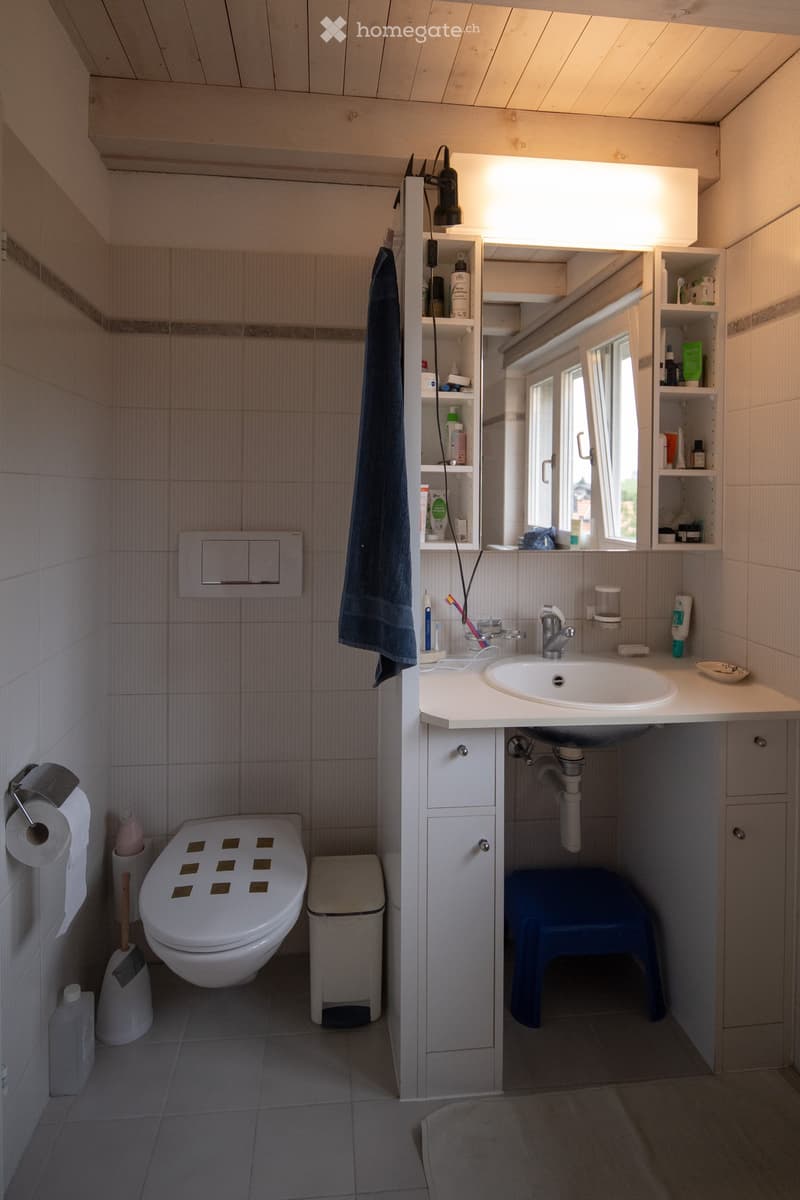 Modern bathroom with a toilet, sink, mirror, and built-in cabinet. Features tiled walls and floor, a window, and a towel hanging on the wall.
