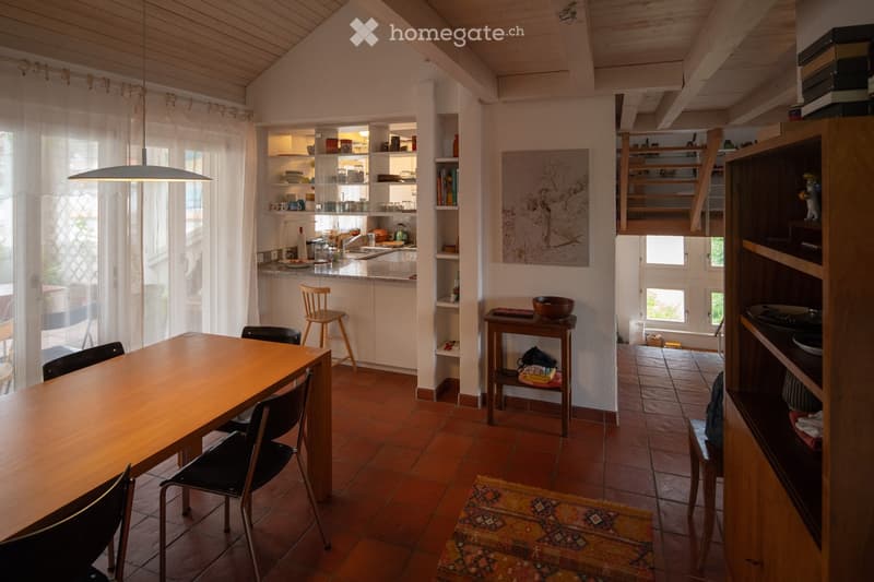 A cozy, open-plan living and dining area with wooden furniture, a kitchen counter, shelves, and a painting on the wall.