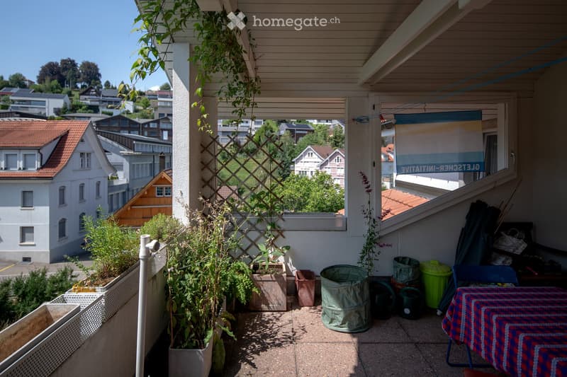 Balcony with potted plants, a small table with a colorful tablecloth, a chair, and various gardening items including a garden hose and trash bins.
