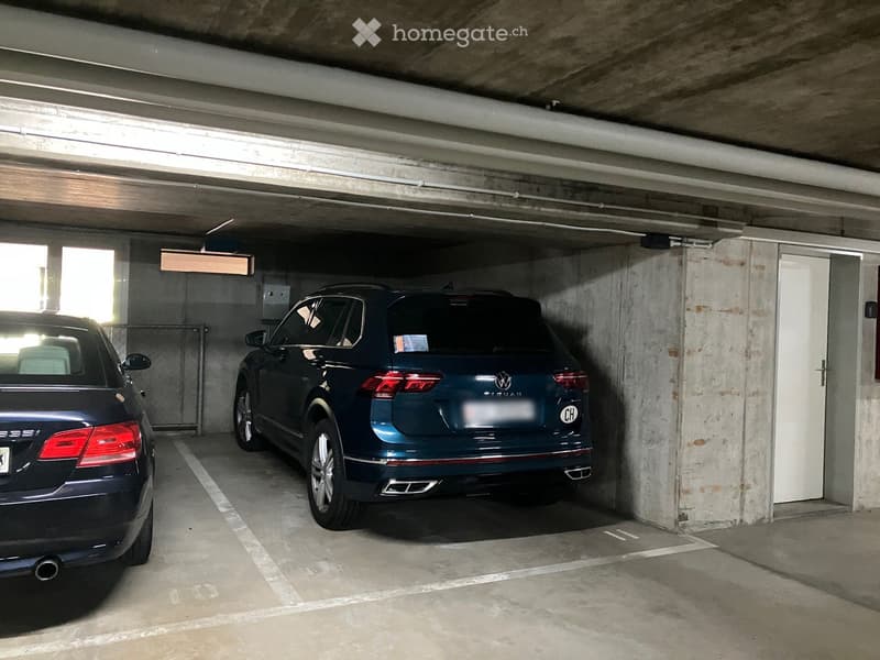Underground parking area with two parked cars, concrete walls, and a white door on the right side.