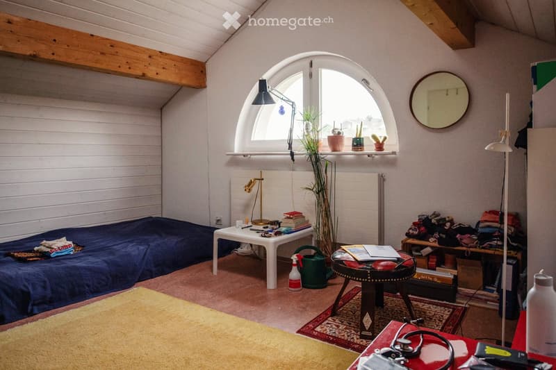 Cozy, well-lit room with a bed, small table, and various decorative items including a mirror, lamps, and plants. The room features wooden beams on the ceiling and a large arched window.