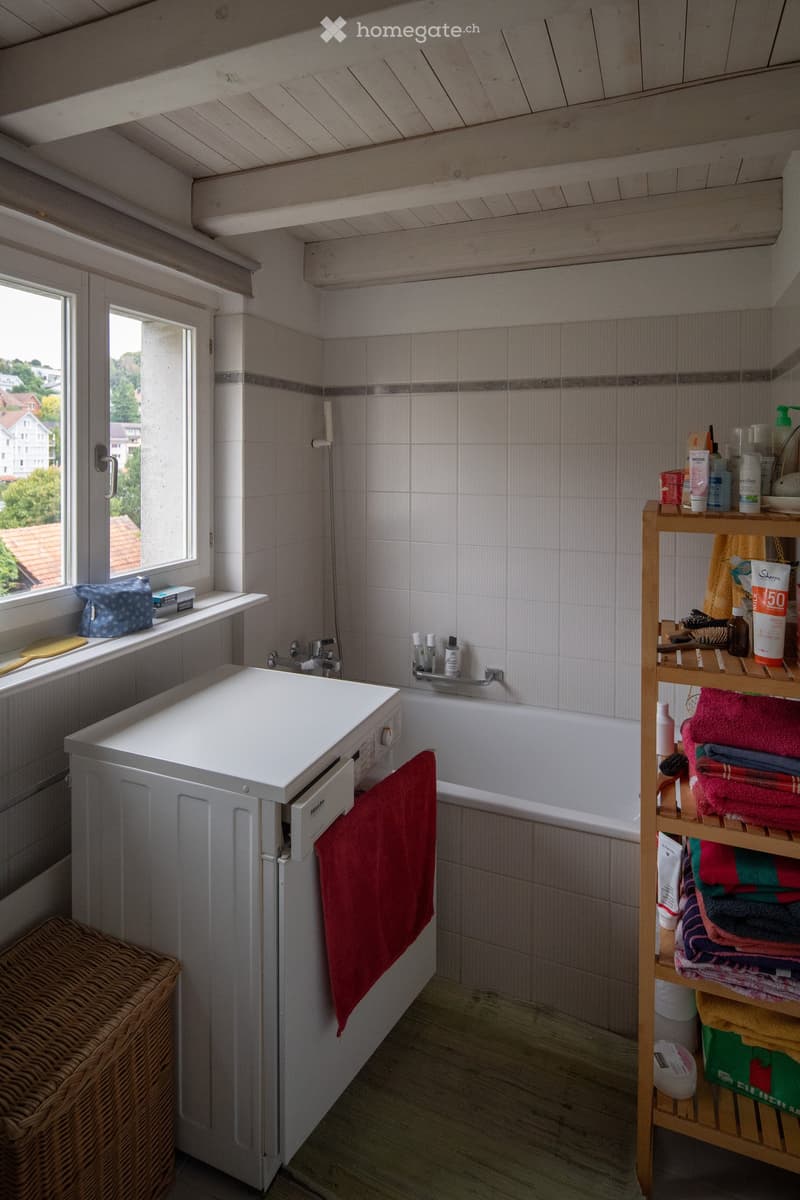 Bathroom with a white sink, bathtub, towel rack, and shelf holding towels and toiletries. A wicker basket is placed on the floor near the window, which offers a view of the outside.