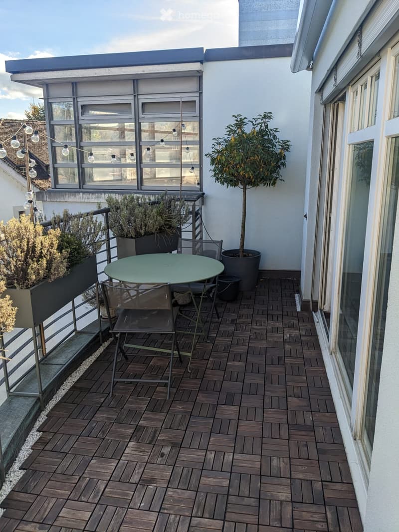 Wooden decked balcony with a round table, chairs, and potted plants, featuring a railing and string lights.