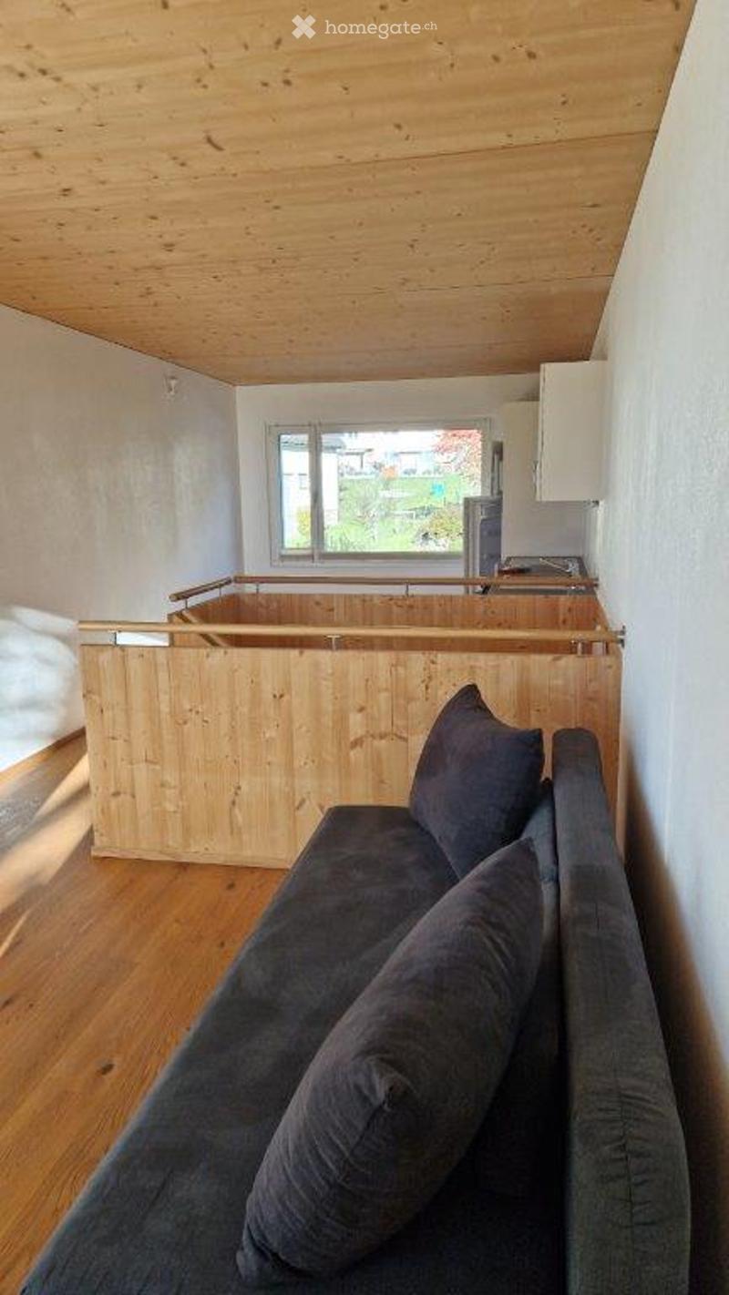 A modern, minimalist living room with a grey sofa, wooden floor, and a large window offering a view of the garden. The room features a wooden ceiling and a small kitchen area in the background.