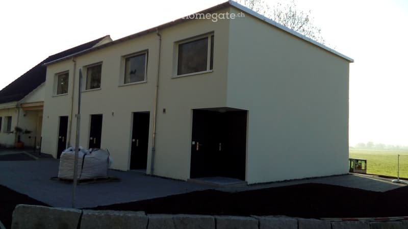 Modern two-story house with white exterior walls, multiple windows, and black doors. The house is situated on a raised ground floor with a paved driveway and a grassy field in the background.