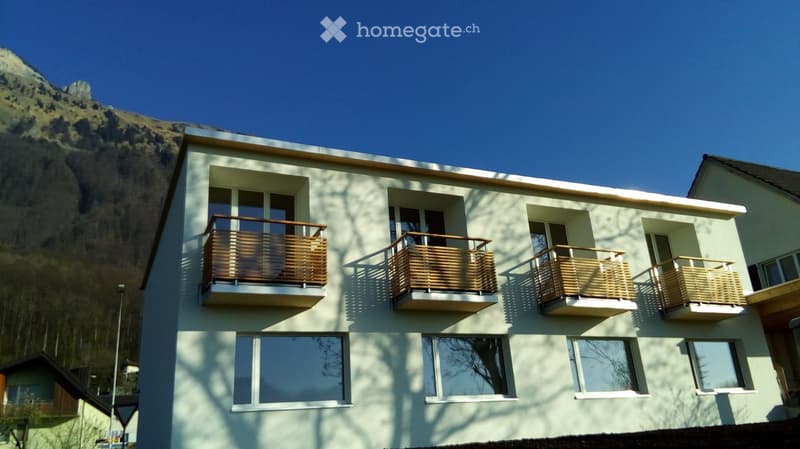 Modern two-story building with light green facade, multiple large windows, and several balconies with wooden railings. The building is situated against a backdrop of a rocky mountain.