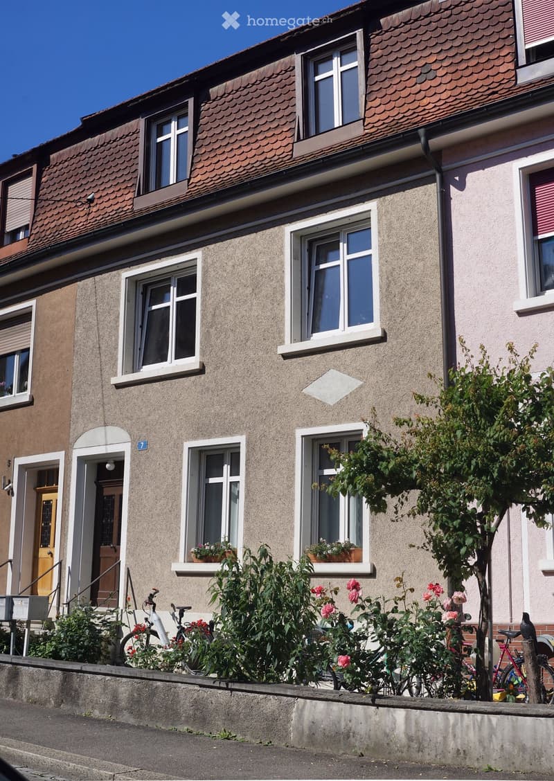 Two-story residential building with beige facade, multiple windows, and a small garden area with bicycles and flowers in front.