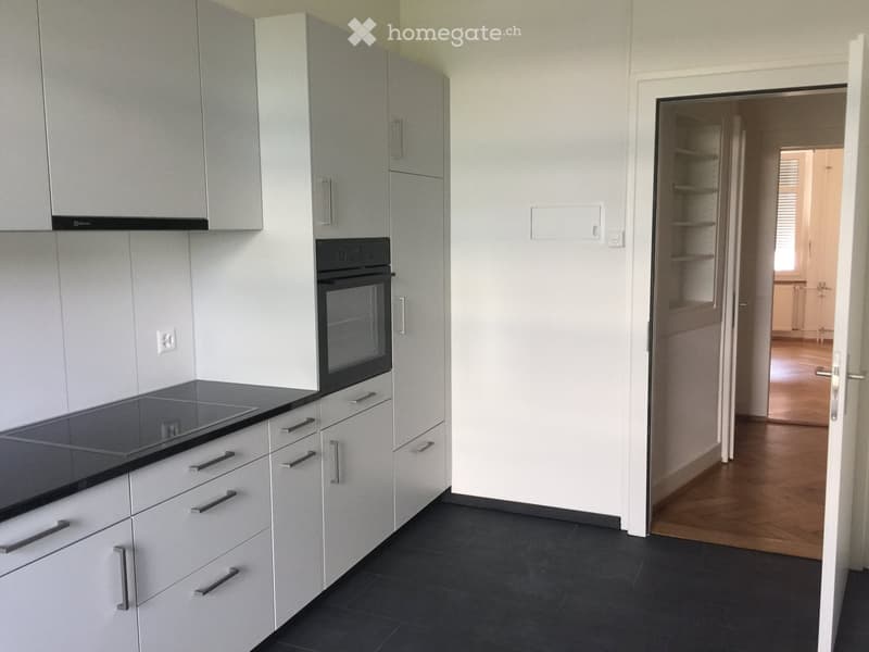 Modern kitchen with white cabinets, black countertop, and built-in oven. Open doorway leads to another room with wooden flooring.