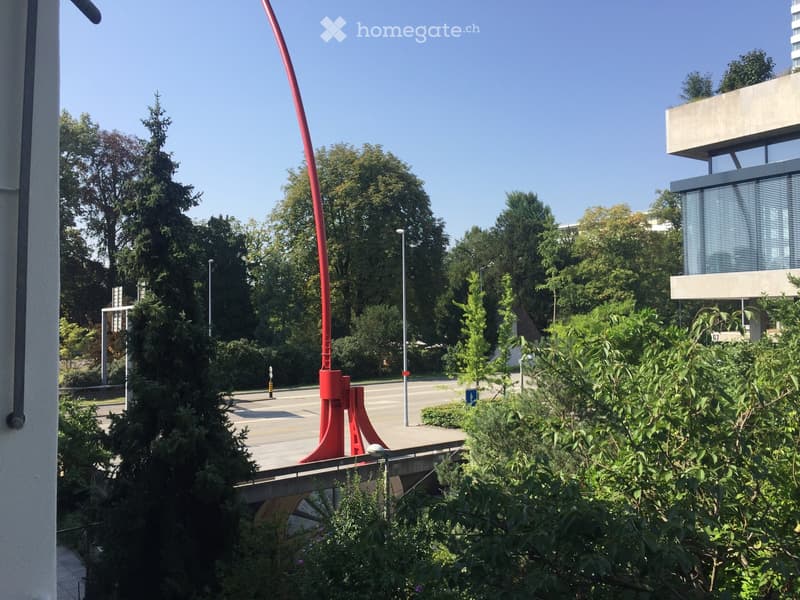 Balcony view showing a landscaped area with trees, a red sculpture, and a modern building in the background.