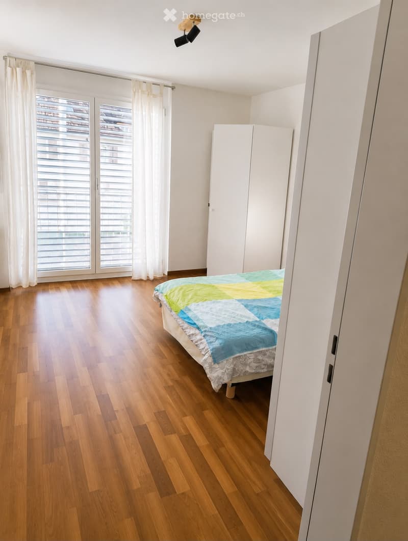 Bright bedroom with wooden flooring, a bed with a colorful quilt, a white wardrobe, and large windows with white curtains.