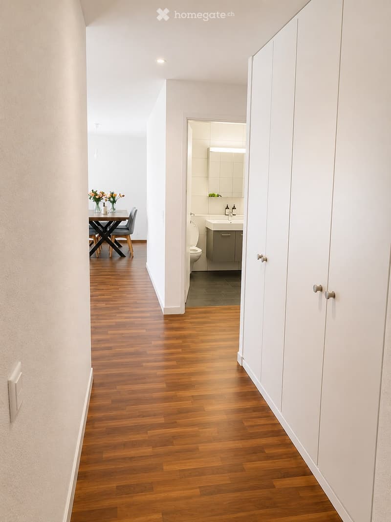 A modern hallway with wooden flooring, white walls, and built-in white cabinets. The hallway leads to a dining area with a small table and chairs, and a bathroom with a sink, toilet, and mirror.