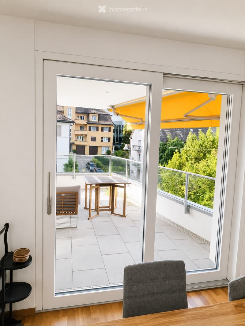 A modern living room with wooden flooring, large sliding glass doors leading to a balcony, and a small seating area on the balcony with a table and chairs.