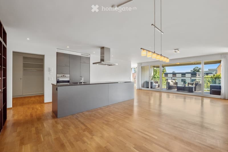 Open-plan kitchen and living area with light wood flooring, modern cabinetry, island, pendant lights, and a view to a furnished living space through large glass doors.